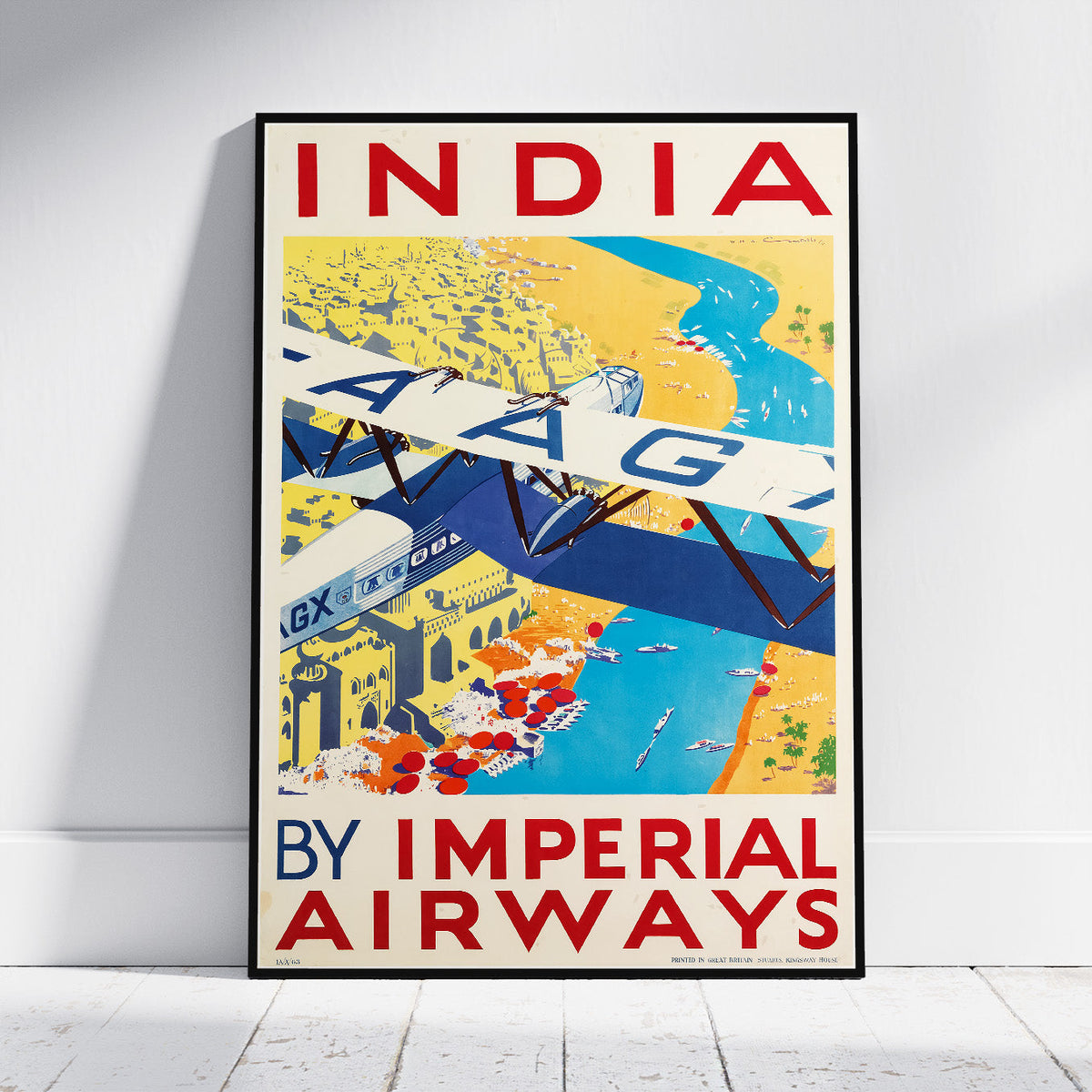 A framed vintage-style travel poster leaning against a white wall on a light floor. The poster reads 'INDIA BY IMPERIAL AIRWAYS' and shows an illustrated airplane over a colorful map scene with yellow land, blue water, and cream background.