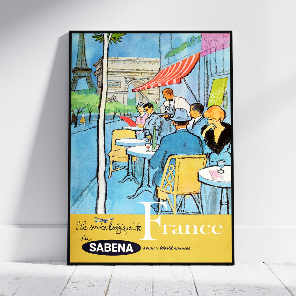 A framed vintage-style travel poster leaning against a white wall on a light floor. The poster depicts a Parisian cafe scene with the Eiffel Tower, Arc de Triomphe, seated figures, and large text reading 'Le plus bel pays du monde France' with 'SABENA' at the bottom.