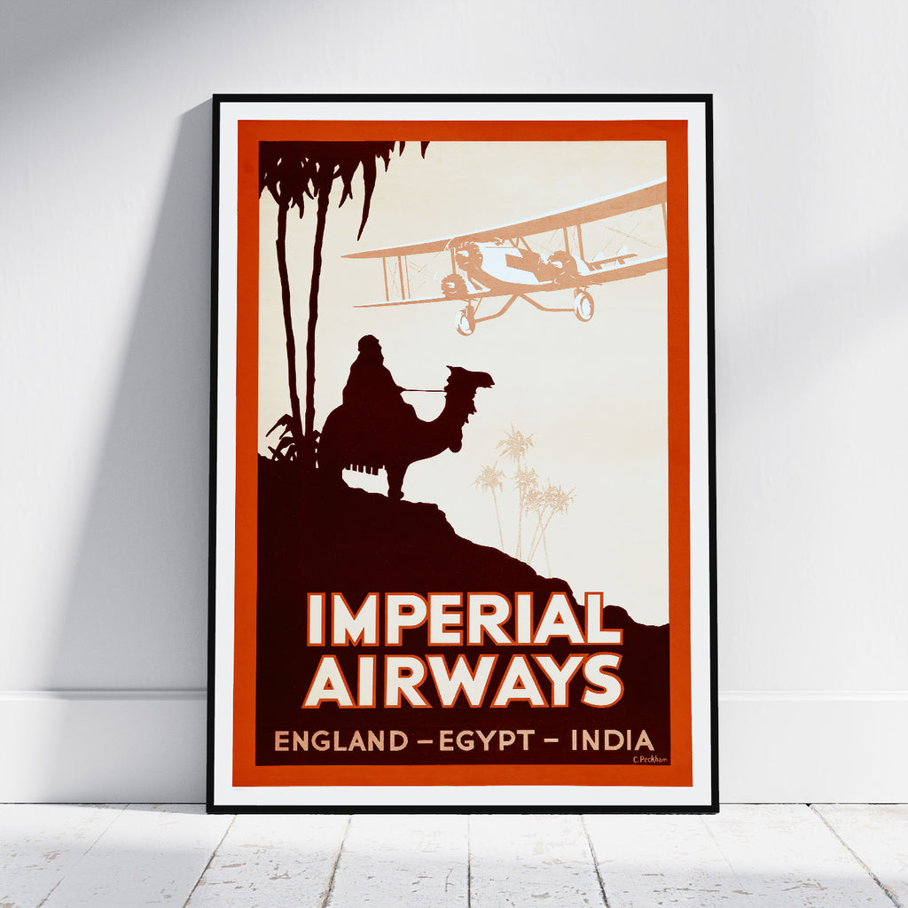 A framed vintage travel poster leaning against a white wall on a light wood floor. The poster features a retro design with the text 'IMPERIAL AIRWAYS' and 'ENGLAND - EGYPT - INDIA', showing a camel rider silhouette, palm trees, and a biplane in warm orange, cream, and dark brown tones.