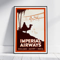 A framed vintage travel poster leaning against a white wall on a light wood floor. The poster features a retro design with the text 'IMPERIAL AIRWAYS' and 'ENGLAND - EGYPT - INDIA', showing a camel rider silhouette, palm trees, and a biplane in warm orange, cream, and dark brown tones.