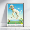A framed vintage-style poster leaning against a white wall on a light wood floor. The poster shows a white lamb jumping in a blue sky with flowers and the text 'Springtime in Switzerland' at the bottom.