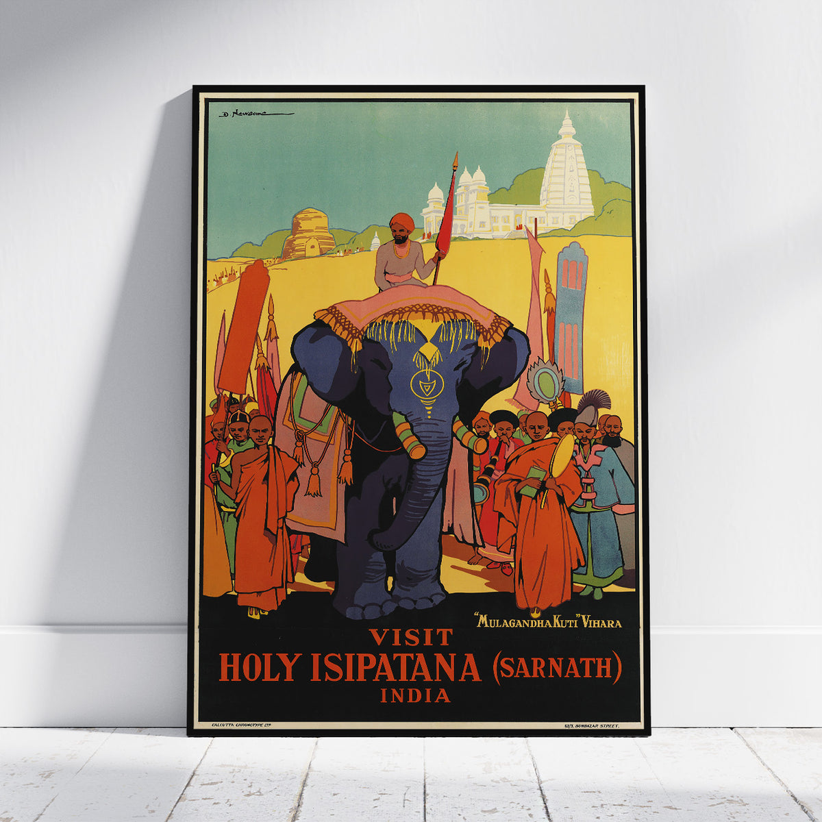 A framed vintage-style travel poster leaning against a white wall on a light floor. The poster depicts a large blue elephant carrying a person on its back, with people in orange robes, a temple in the background, and the text 'VISIT HOLY ISIPATAN (SARNATH) INDIA' at the bottom.