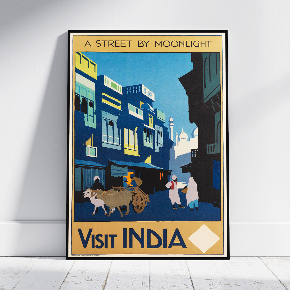 A framed travel poster leaning against a white wall on a light wood floor. The poster features an illustration of an Indian street at night with blue buildings, a bullock cart, and people, with the text 'A STREET BY MOONLIGHT' at the top and 'VISIT INDIA' at the bottom.