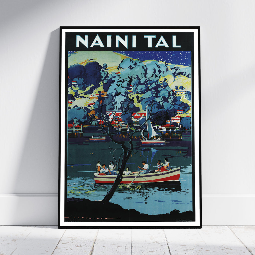 A framed vintage-style travel poster leaning against a white wall on a light floor. The poster reads "NAINI TAL" at the top and depicts a scenic lake or river scene with a boat, trees, buildings, and a starry sky in blue, teal, cream, and red tones.