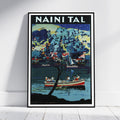 A framed vintage-style travel poster leaning against a white wall on a light floor. The poster reads "NAINI TAL" at the top and depicts a scenic lake or river scene with a boat, trees, buildings, and a starry sky in blue, teal, cream, and red tones.