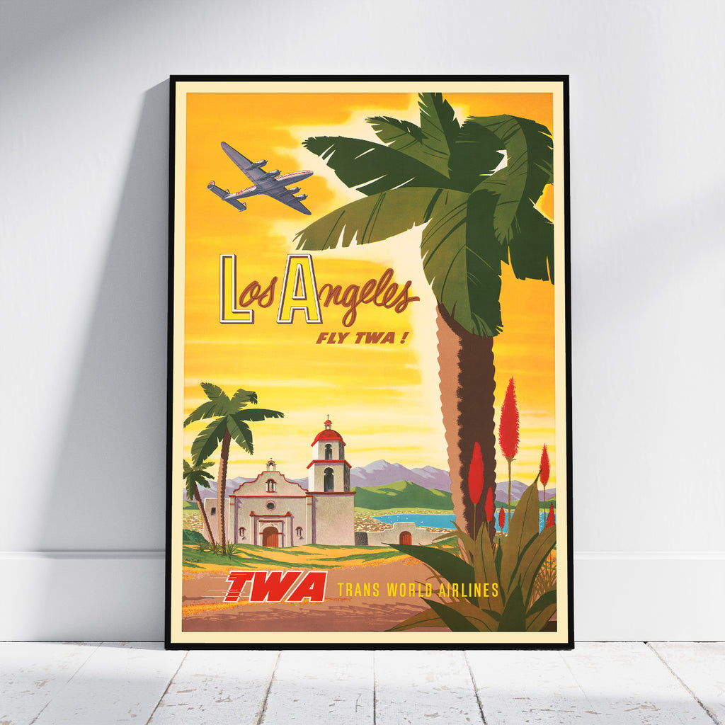 A framed vintage-style Los Angeles travel poster leaning against a white wall on a light wood floor. The poster features a yellow-orange sky, a large palm tree, an airplane, a church, and the text 'Los Angeles Fly TWA' and 'TWA Trans World Airlines'.