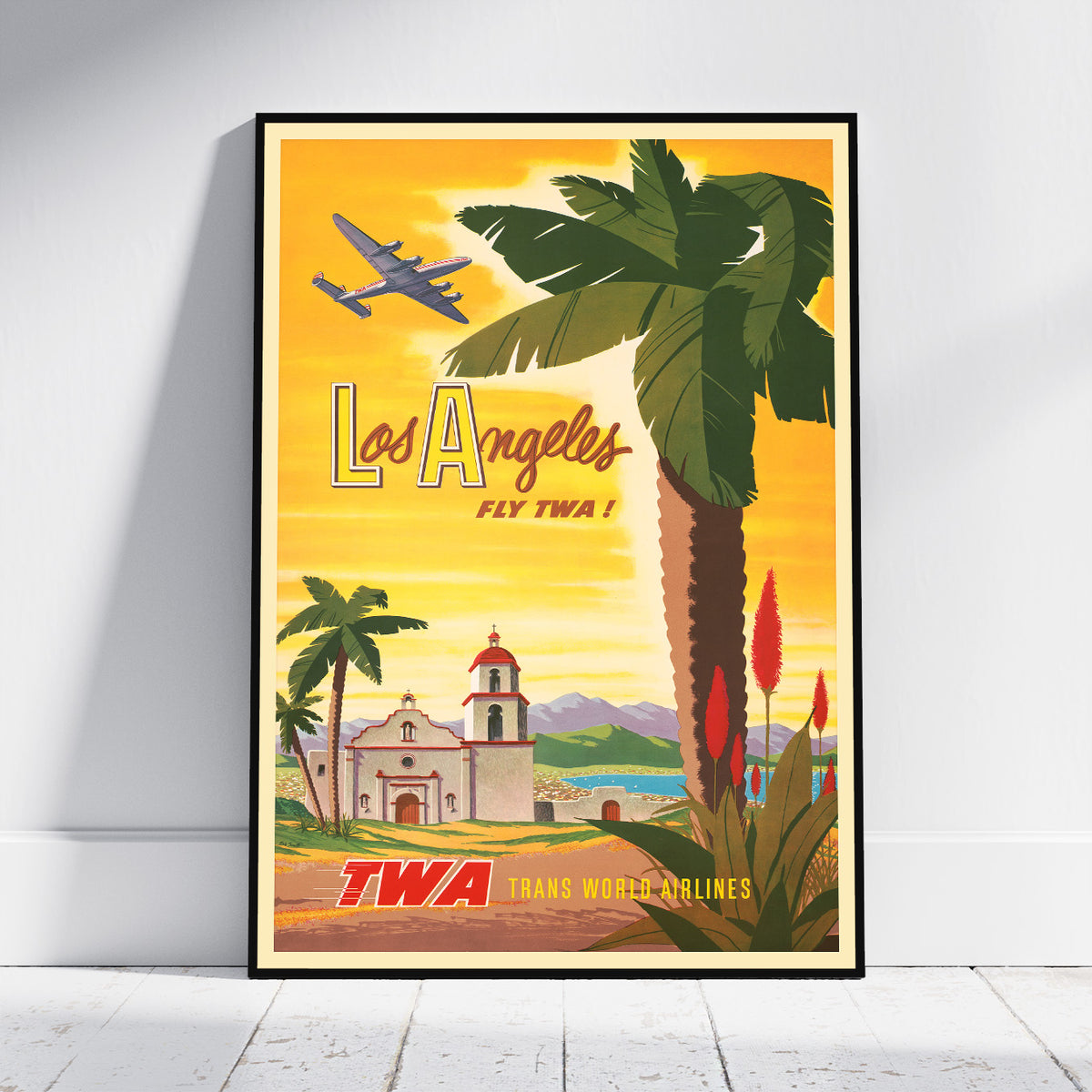 A framed vintage-style Los Angeles travel poster leaning against a white wall on a light wood floor. The poster features a yellow-orange sky, a large palm tree, an airplane, a church, and the text 'Los Angeles Fly TWA' and 'TWA Trans World Airlines'.