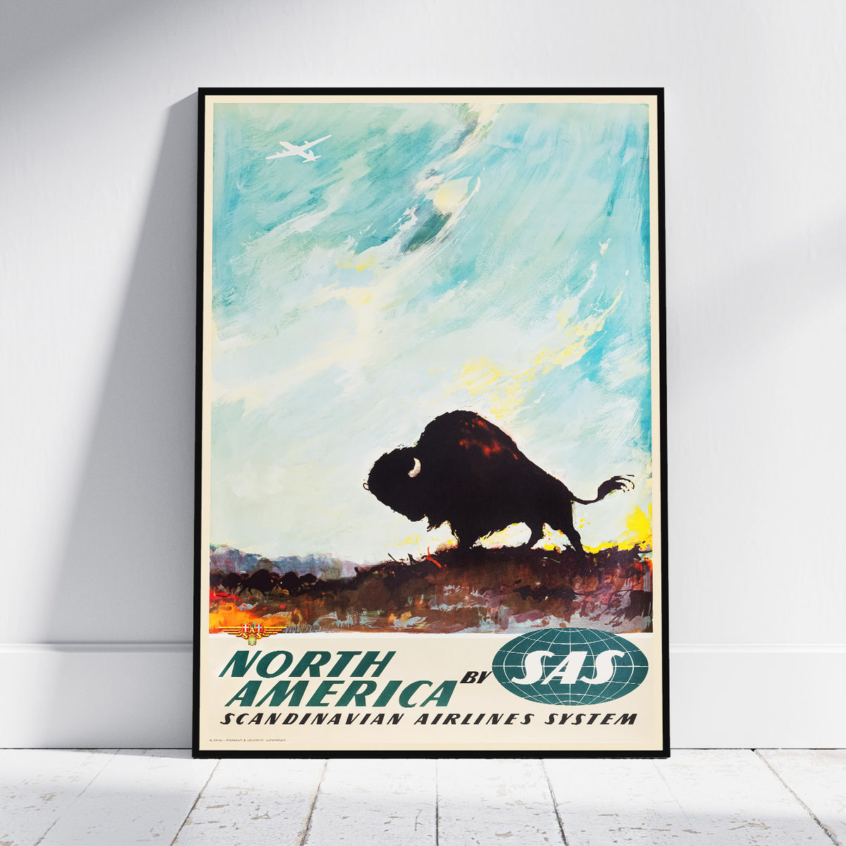 A framed vintage-style travel poster leaning against a white wall on a light wood floor. The poster shows a bison silhouette against a blue sky with clouds, and the text 'NORTH AMERICA by SAS' with 'SCANDINAVIAN AIRLINES SYSTEM' below.