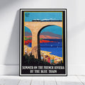 A framed vintage-style travel poster leaning against a white wall on a light wood floor. The poster depicts a blue train traveling under a large stone bridge over a coastal landscape, with the text 'SUMMER ON THE FRENCH RIVIERA BY THE BLUE TRAIN' at the bottom.