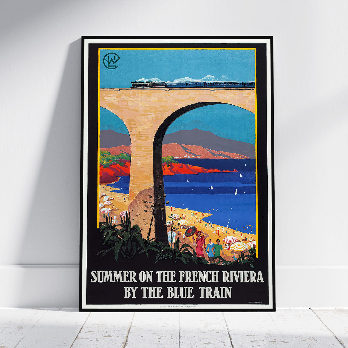 A framed vintage-style travel poster leaning against a white wall on a light wood floor. The poster depicts a blue train traveling under a large stone bridge over a coastal landscape, with the text 'SUMMER ON THE FRENCH RIVIERA BY THE BLUE TRAIN' at the bottom.