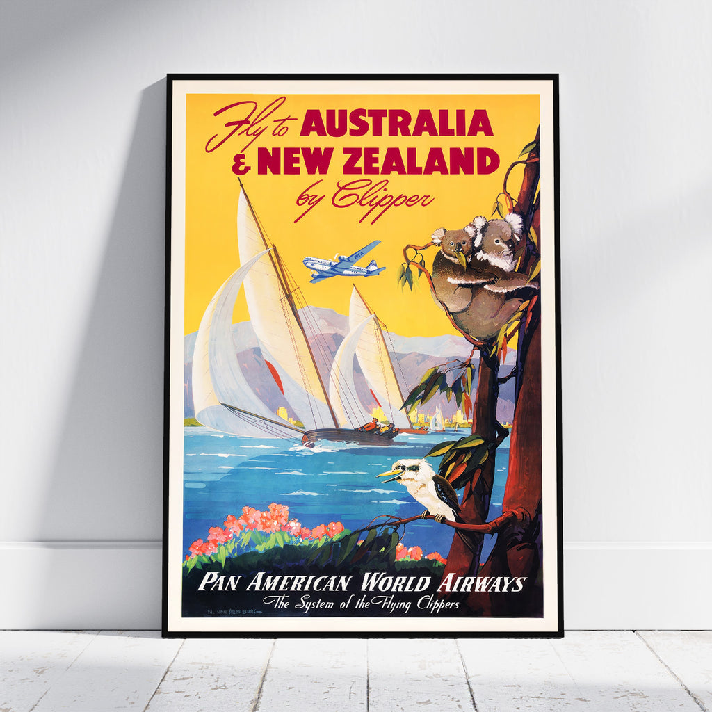 A framed vintage-style travel poster leaning against a white wall on a light wood floor. The poster features a yellow sky, sailboats on blue water, an airplane, a koala in a tree, and text reading 'Fly to Australia & New Zealand by Clipper' and 'Pan American World Airways.