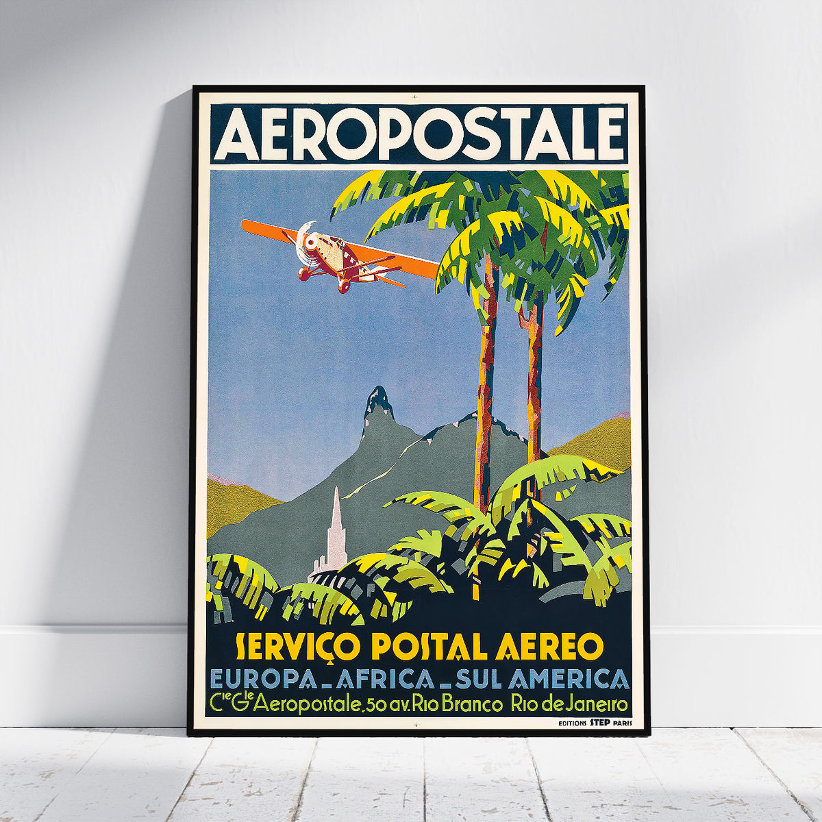A framed vintage-style travel poster leaning against a white wall on a light floor. The poster reads 'AEROPOSTALE' at the top and features an illustration of a biplane flying over tropical palm trees and a mountain landscape, with yellow and blue typography near the bottom.