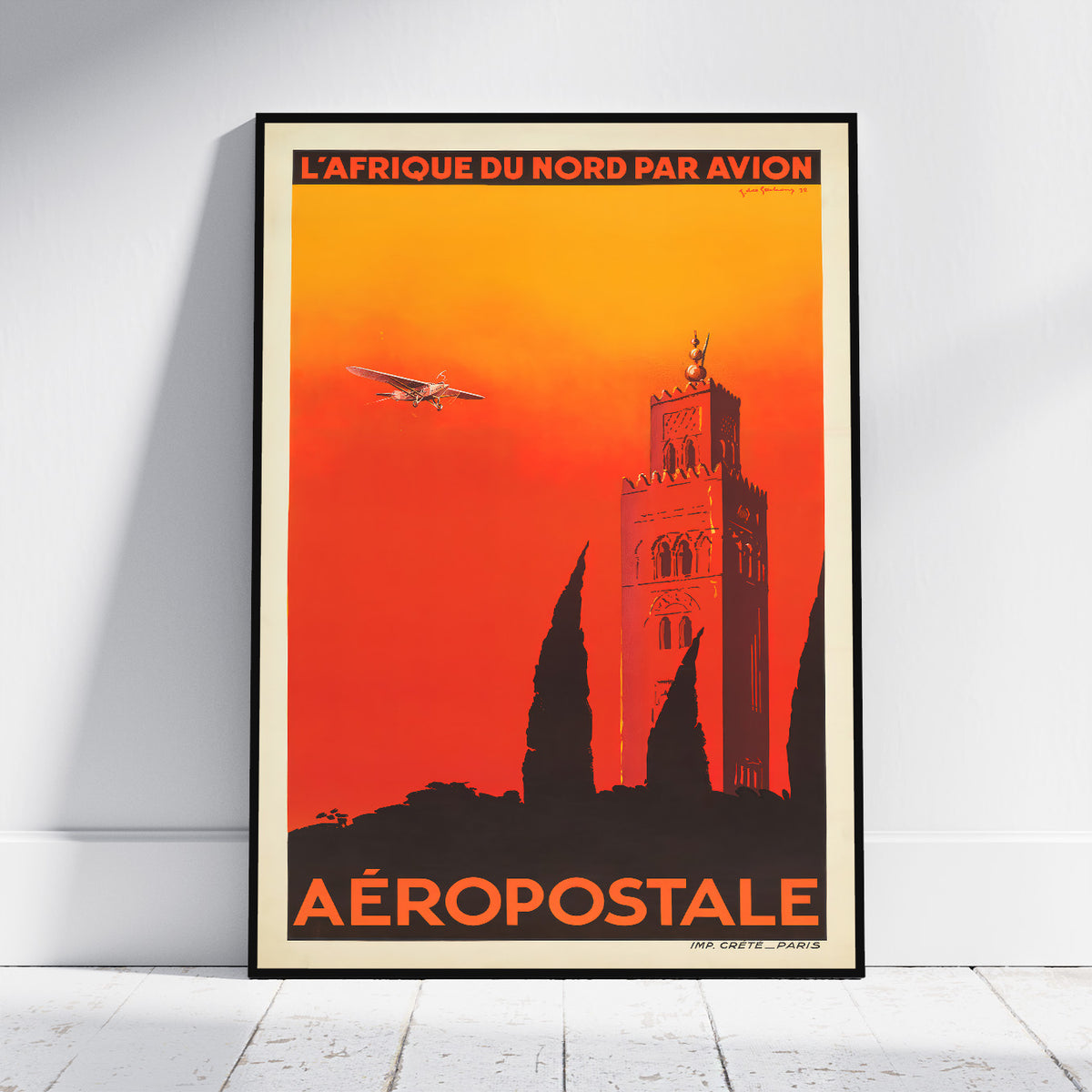 A framed vintage-style travel poster leaning against a white wall on a light floor. The poster reads 'L'AFRIQUE DU NORD PAR AVION' at the top and 'AÉROPOSTALE' at the bottom, with an orange-to-red sky, a tower silhouette, and an airplane illustration.