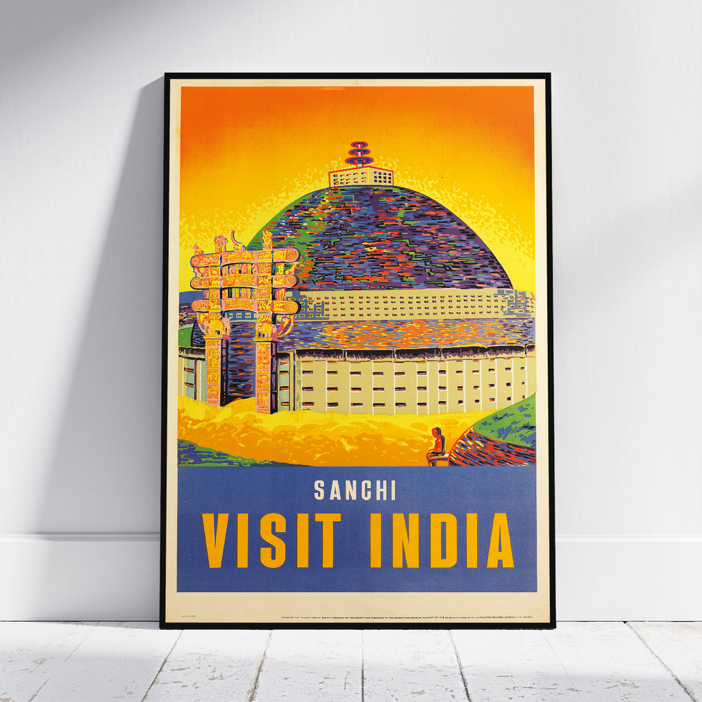 A framed travel poster of Sanchi, India, leaning against a white wall on a light wood floor. The poster features a vibrant, textured illustration of the Sanchi Stupa under a bright yellow and orange sky, with a blue banner at the bottom reading 'SANCHI VISIT INDIA'.