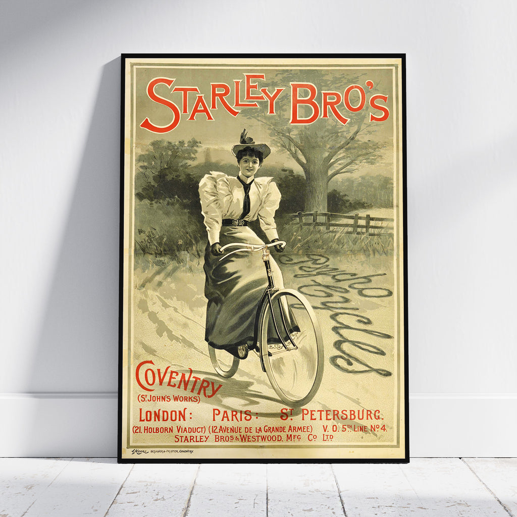 A framed vintage-style poster leaning against a white wall on a light floor. The poster reads 'Starley Bro's' and shows an illustrated woman riding a bicycle in a sepia landscape, with red and black text and a black frame around the print.