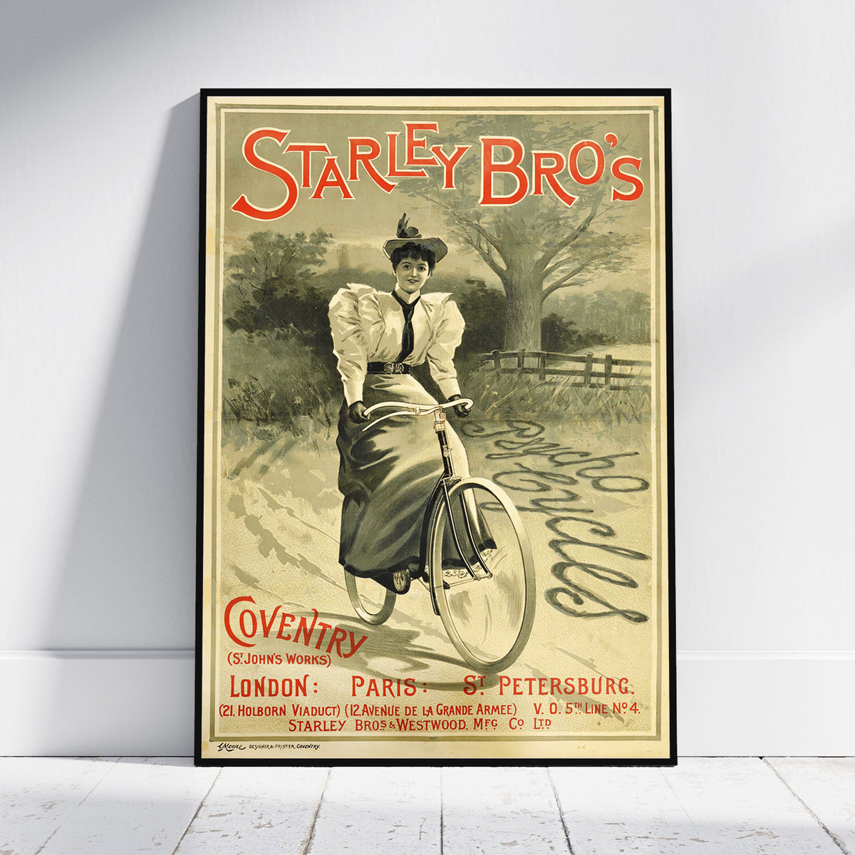 A framed vintage-style poster leaning against a white wall on a light floor. The poster reads 'Starley Bro's' and shows an illustrated woman riding a bicycle in a sepia landscape, with red and black text and a black frame around the print.