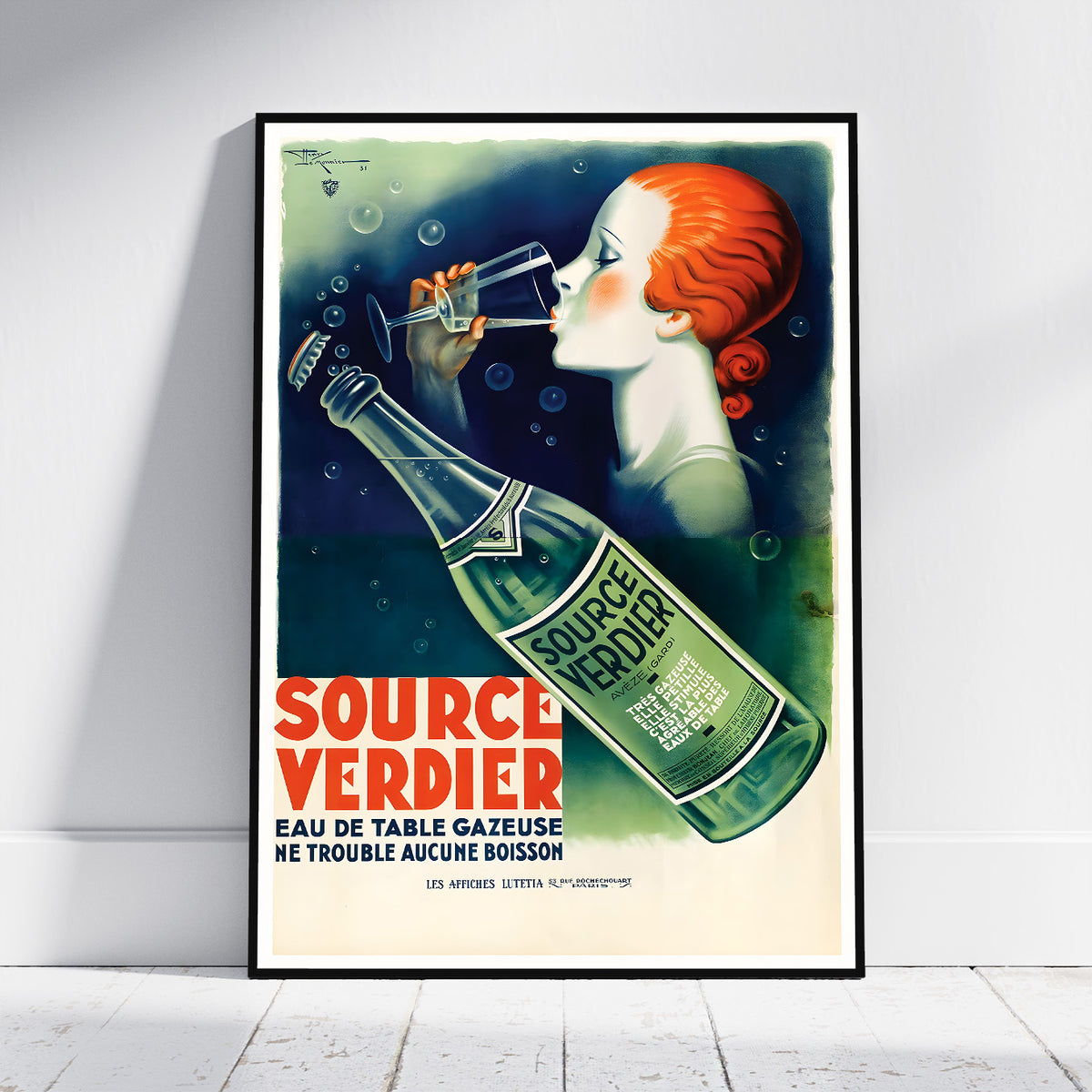 A framed vintage-style beer advertisement poster leaning against a white wall on a light floor. The artwork shows a woman with red hair drinking from a glass bottle, with the text 'SOURCE VERDIER' in large red letters and 'EAU DE TABLE GAZEUSE' below.