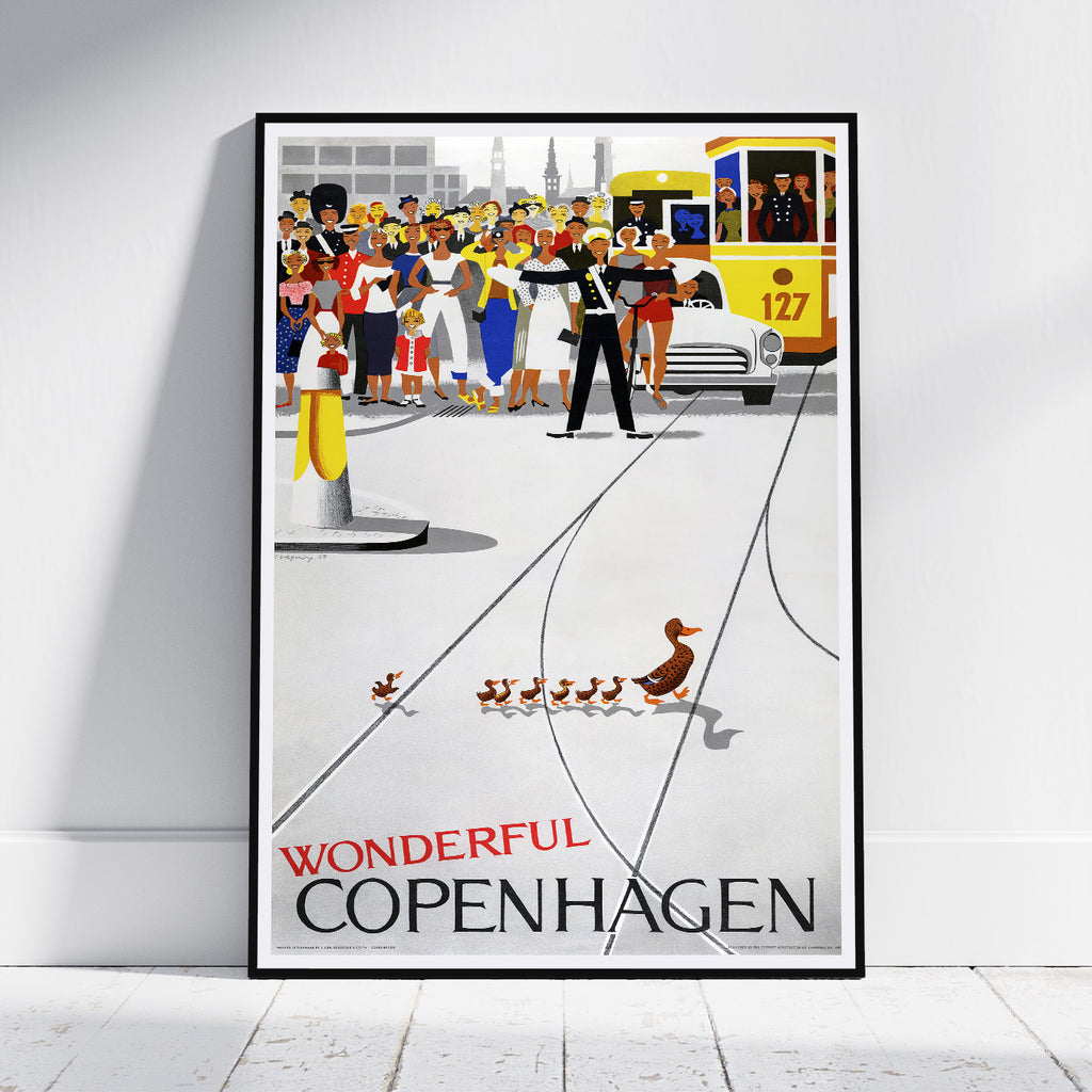 A framed vintage-style travel poster leaning against a white wall on a light floor. The poster depicts a busy Copenhagen street scene with a yellow tram, pedestrians, and a duck family, with the text 'WONDERFUL COPENHAGEN' at the bottom.