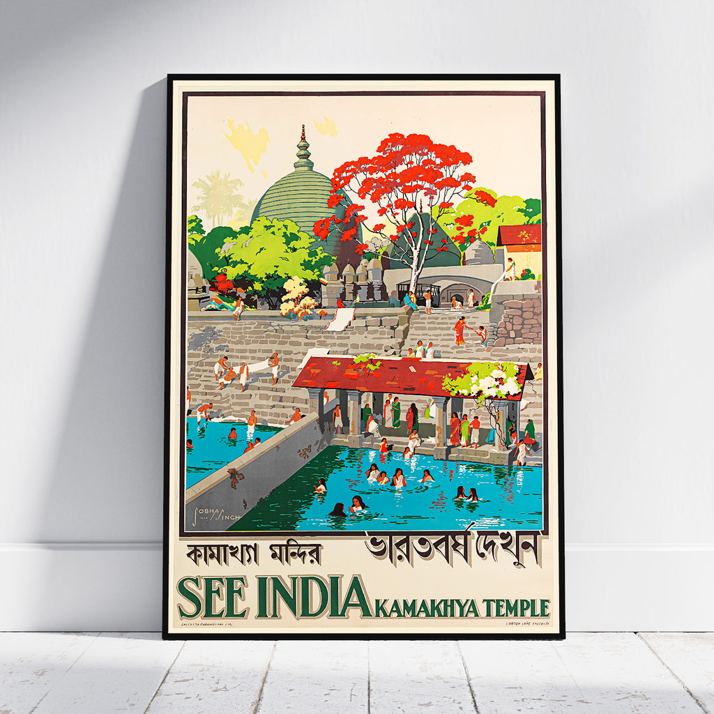 A framed travel poster of Kamakhiya Temple in Rameswaram, India, leaning against a white wall on a light wood floor. The poster features a retro-style illustration with a green stupa, a red-leafed tree, and people swimming in the blue water of the temple pool, with text reading 'SEE INDIA KAMAKHIYA TEMPLE'.