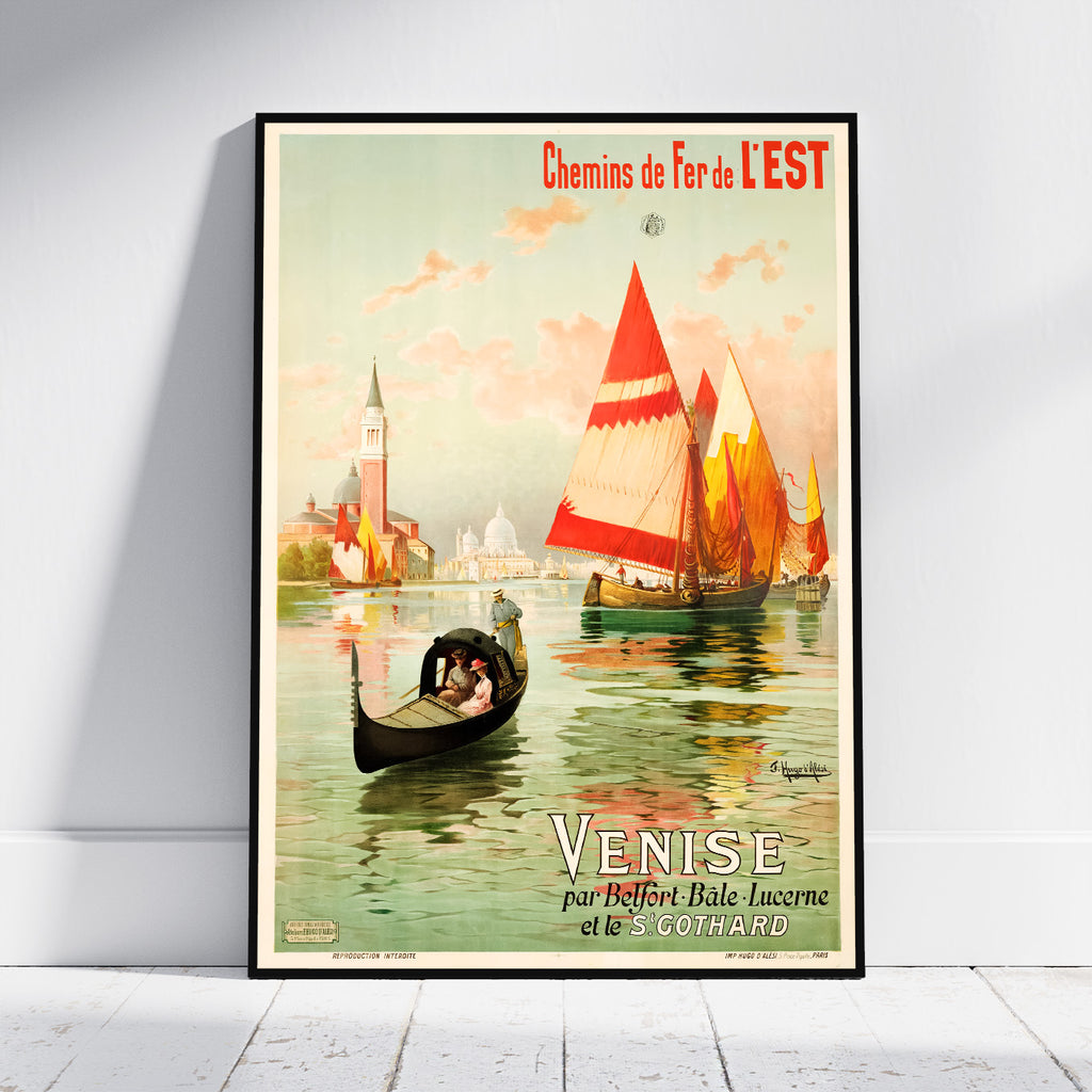 A framed vintage travel poster of Venice, France, leaning against a white wall on a light wood floor. The poster features a gondola with a passenger and sailboats on the water under a pale sky, with French text reading 'Chemin de Fer de L'EST' and 'VENISE'.