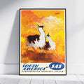 A framed vintage-style travel poster leaning against a white wall on a light floor. The artwork shows two llama-like animals in a warm orange and yellow landscape with mountains, and the text 'SOUTH AMERICA by SAS' at the bottom.