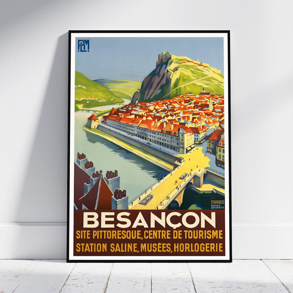 A framed vintage-style travel poster of Besancon, France, leaning against a white wall on a light wood floor. The poster features a colorful illustration of a river town with red-roofed buildings, green hills, and a bridge, with the text 'BESANCON' and additional French text at the bottom.