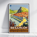 A framed vintage-style travel poster of Besancon, France, leaning against a white wall on a light wood floor. The poster features a colorful illustration of a river town with red-roofed buildings, green hills, and a bridge, with the text 'BESANCON' and additional French text at the bottom.