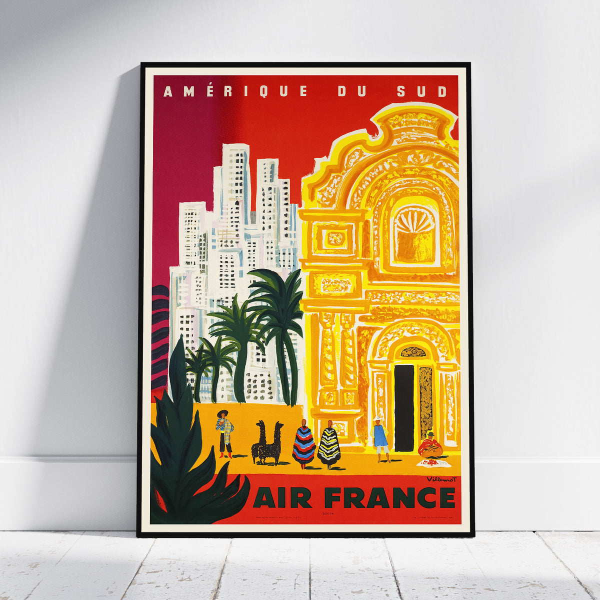 A framed vintage-style travel poster leaning against a white wall on a light wood floor. The poster features bold red, yellow, and orange artwork with the text “AMÉRIQUE DU SUD” at the top and “AIR FRANCE” at the bottom, depicting a stylized cityscape and ornate building.