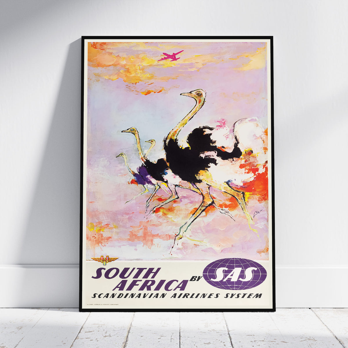 A framed vintage-style travel poster leaning against a white wall on a light floor. The artwork shows ostriches and a pink airplane over a pastel sky, with the text 'SOUTH AFRICA by SAS' and 'SCANDINAVIAN AIRLINES SYSTEM' at the bottom.