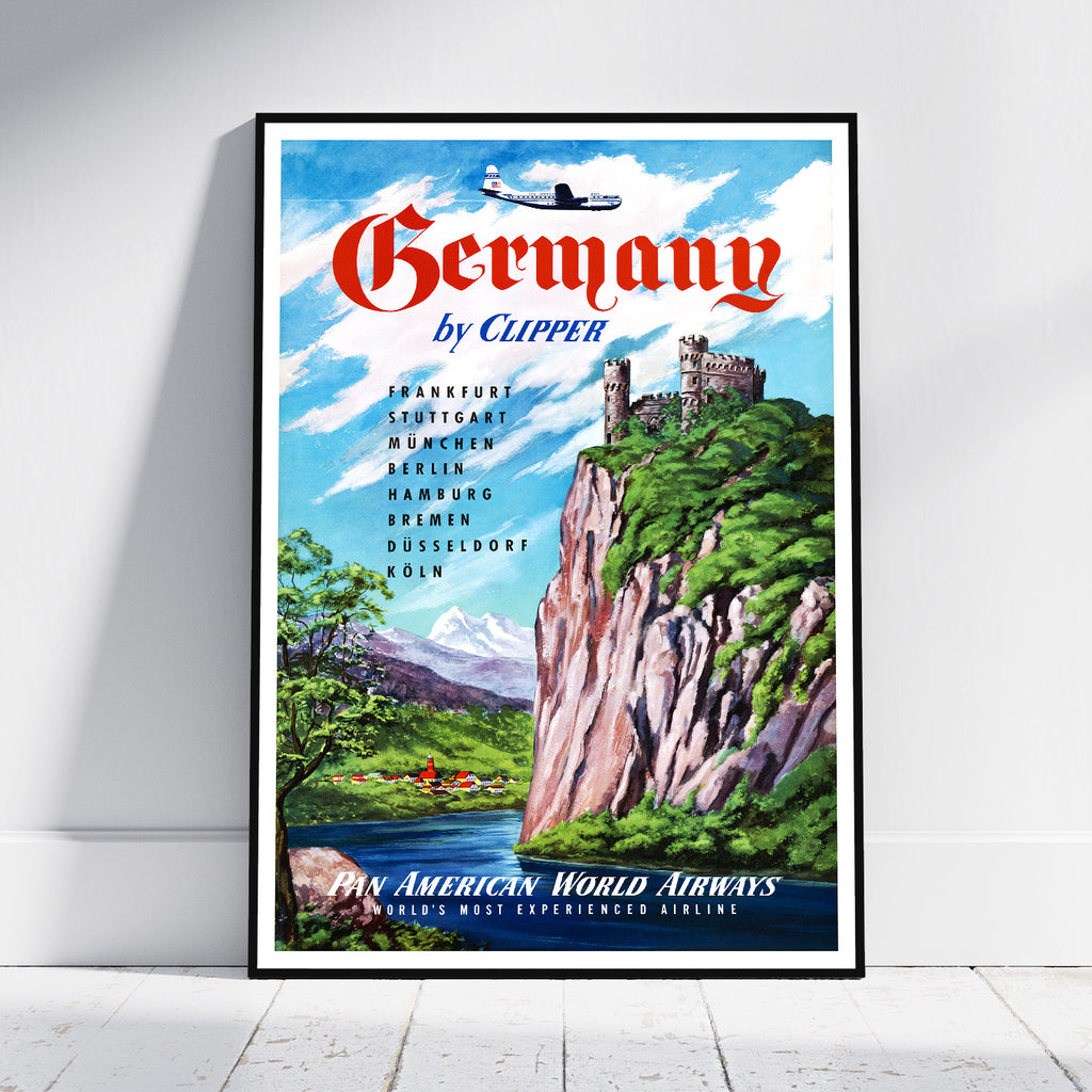 A framed vintage-style travel poster leaning against a white wall on a light floor. The poster reads 'Germany by Clipper' with red lettering, a blue sky, green cliffs, mountains, and a small airplane illustration.