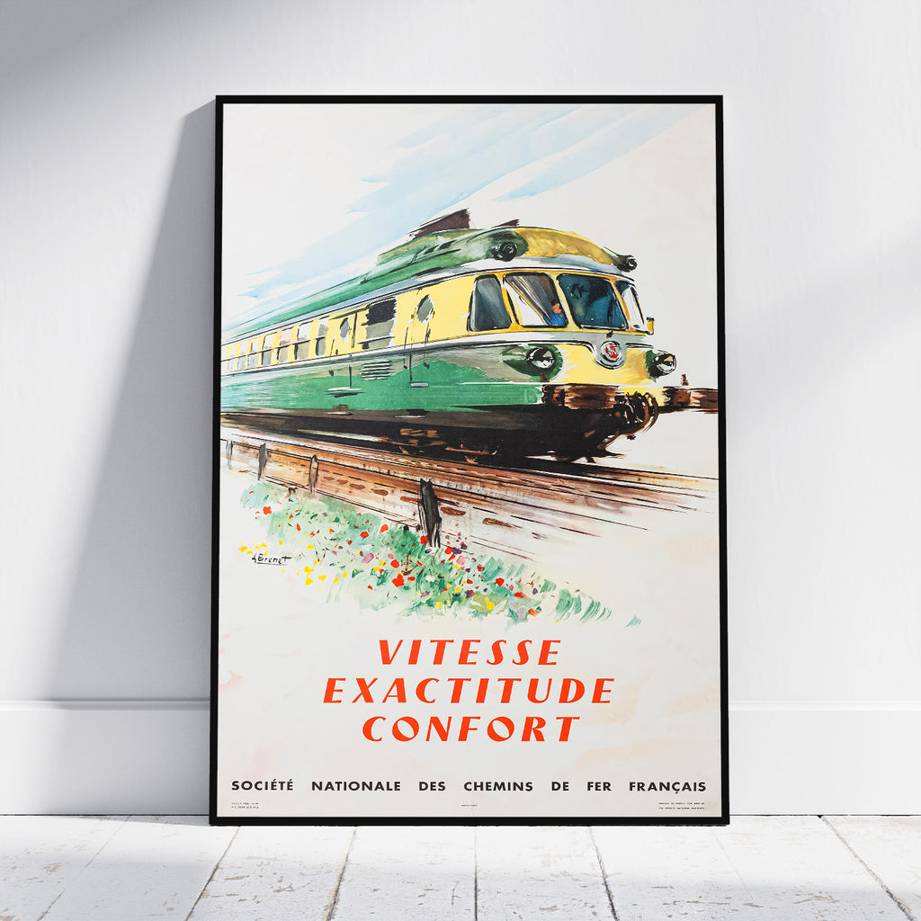 A framed vintage French train poster leaning against a white wall on a light floor. The poster shows a green and yellow train on tracks with the text 'VITESSE EXACTITUDE CONFORT' and smaller text at the bottom reading 'SOCIETE NATIONALE DES CHEMINS DE FER FRANCAIS'.