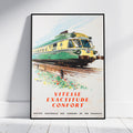 A framed vintage French train poster leaning against a white wall on a light floor. The poster shows a green and yellow train on tracks with the text 'VITESSE EXACTITUDE CONFORT' and smaller text at the bottom reading 'SOCIETE NATIONALE DES CHEMINS DE FER FRANCAIS'.