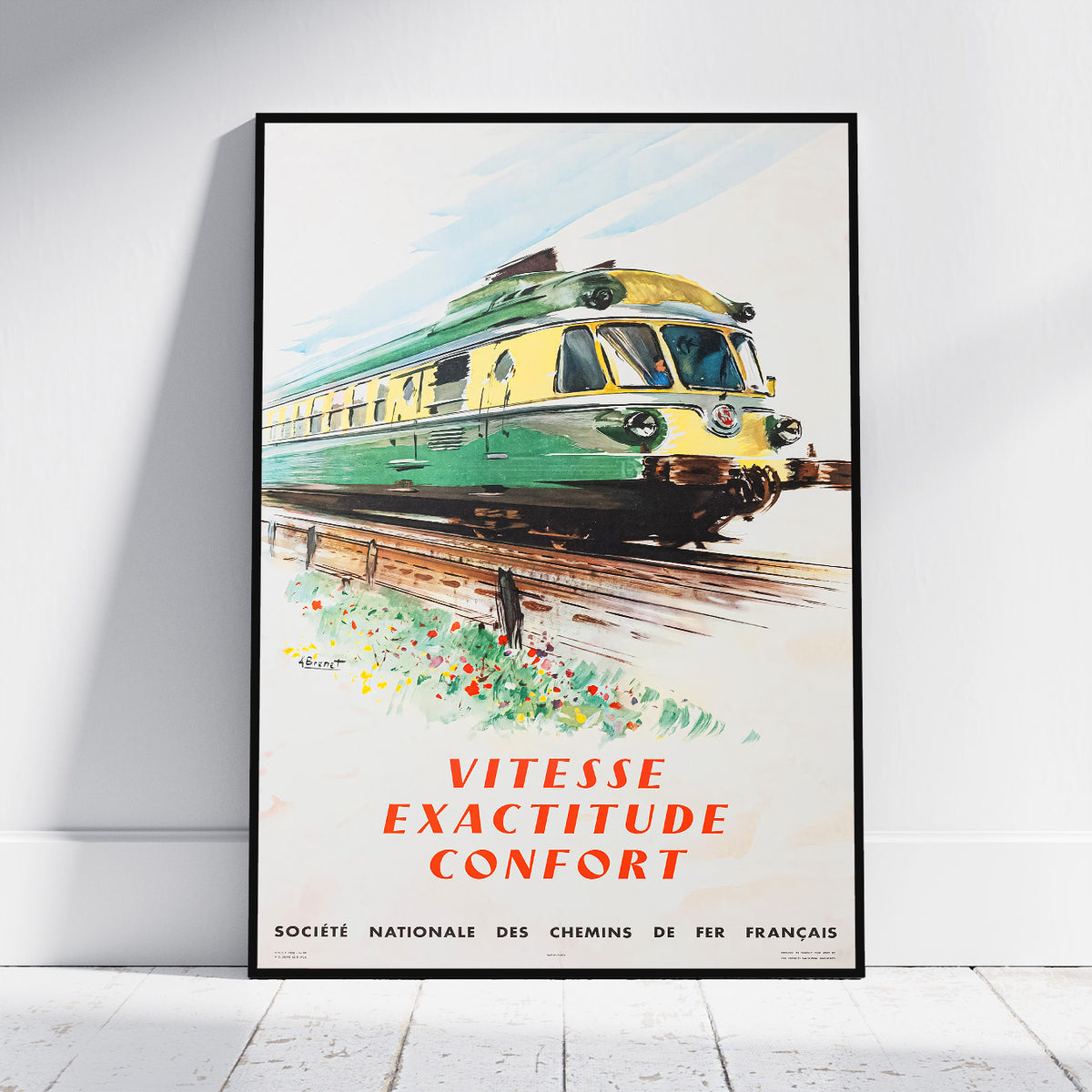A framed vintage French train poster leaning against a white wall on a light floor. The poster shows a green and yellow train on tracks with the text 'VITESSE EXACTITUDE CONFORT' and smaller text at the bottom reading 'SOCIETE NATIONALE DES CHEMINS DE FER FRANCAIS'.