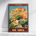 A framed travel poster of Jodhpur, India, leaning against a white wall on a light wood floor. The poster features a vintage-style illustration of the orange-red Jodhpur Fort and surrounding desert landscape with green trees and buildings, framed with red borders at the top and bottom containing the text 'JODHPUR' and 'SEE INDIA'.