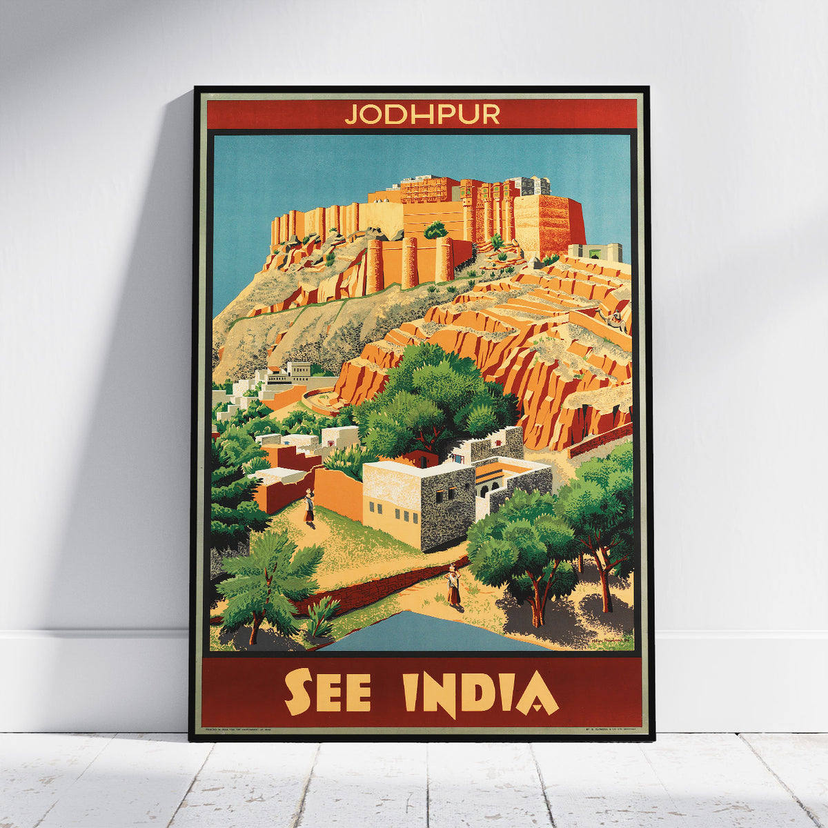 A framed travel poster of Jodhpur, India, leaning against a white wall on a light wood floor. The poster features a vintage-style illustration of the orange-red Jodhpur Fort and surrounding desert landscape with green trees and buildings, framed with red borders at the top and bottom containing the text 'JODHPUR' and 'SEE INDIA'.
