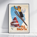 A framed vintage-style travel poster leaning against a white wall on a light wood floor. The poster shows a skier above a red mountain peak with the text 'VAL DAosta' in large orange letters.