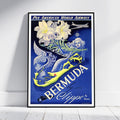 A framed vintage-style travel poster for Pan American World Airways featuring Bermuda by Clipper, leaning against a white wall on a light floor. The artwork shows a blue background with large white lilies, a yellow-and-blue mermaid, an airplane, and bold white typography.