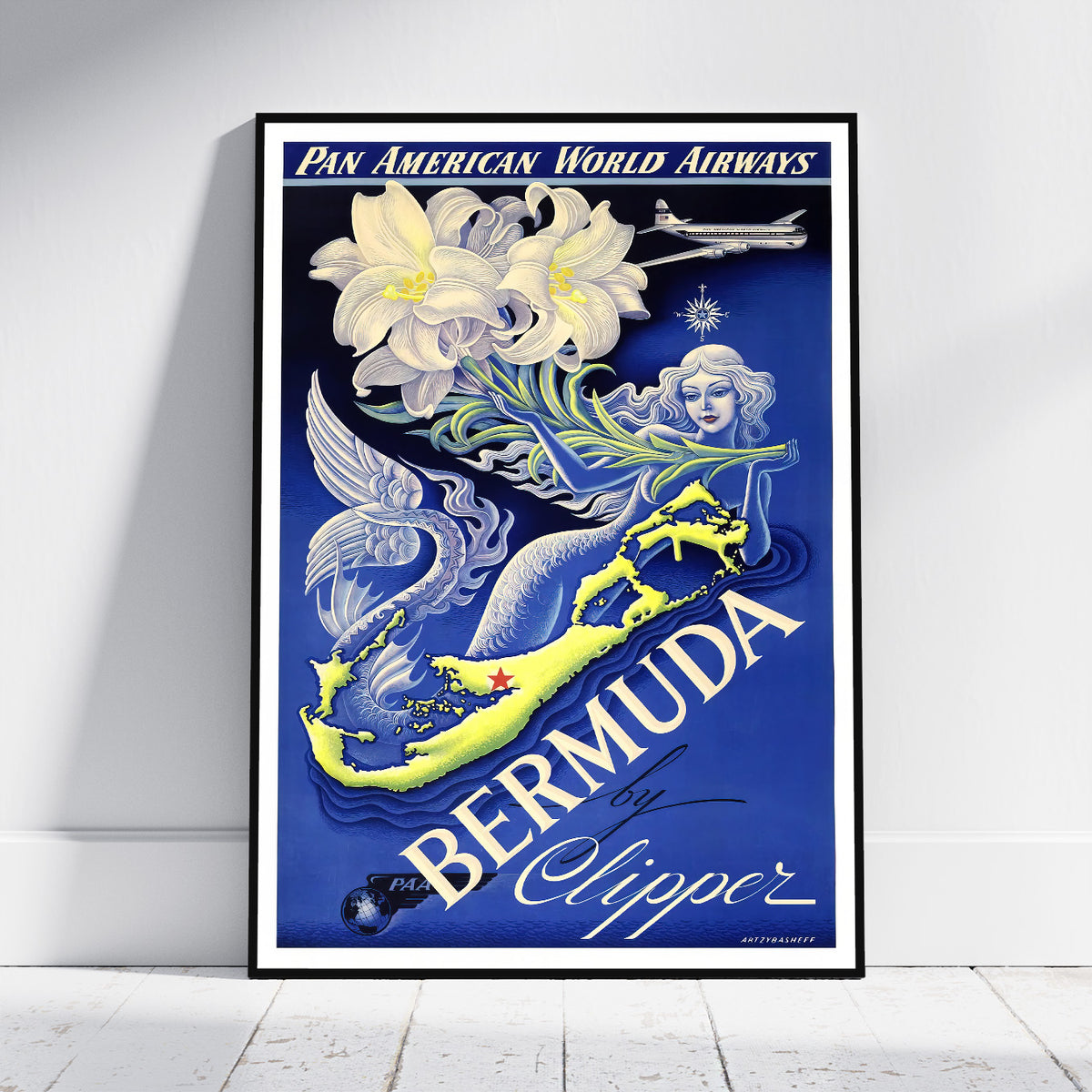 A framed vintage-style travel poster for Pan American World Airways featuring Bermuda by Clipper, leaning against a white wall on a light floor. The artwork shows a blue background with large white lilies, a yellow-and-blue mermaid, an airplane, and bold white typography.