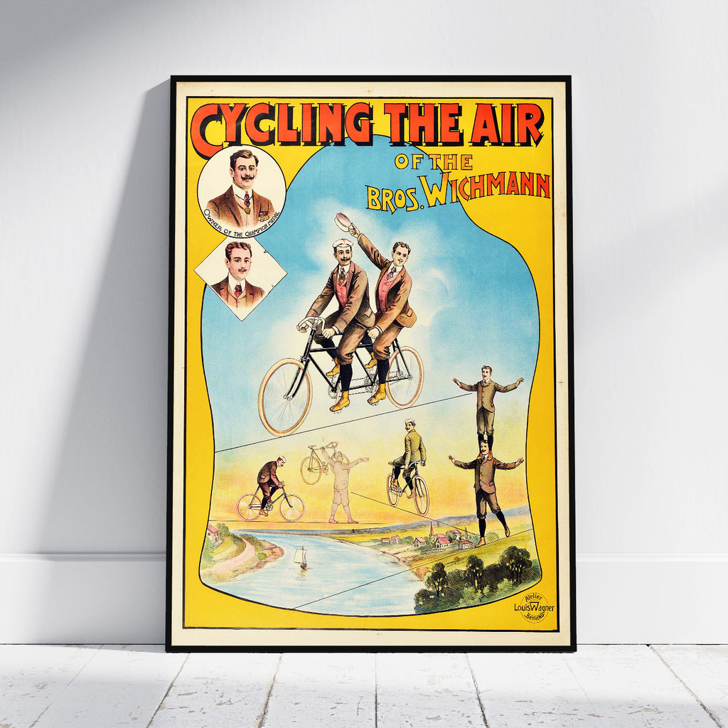 A framed vintage-style cycling poster leaning against a white wall on a light floor. The poster has a yellow border, red title text reading 'CYCLING THE AIR', and an illustrated scene with multiple cyclists and riders, plus portrait insets at the upper left.