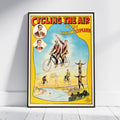 A framed vintage-style cycling poster leaning against a white wall on a light floor. The poster has a yellow border, red title text reading 'CYCLING THE AIR', and an illustrated scene with multiple cyclists and riders, plus portrait insets at the upper left.