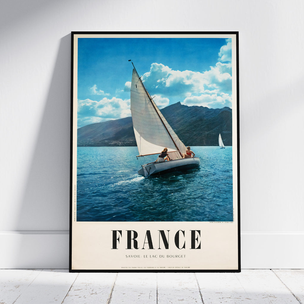 A framed vintage-style travel poster leaning against a white wall on a light floor. The poster shows a sailboat on blue water with mountains and a cloudy sky, and the word FRANCE in large letters at the bottom.