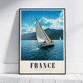 A framed vintage-style travel poster leaning against a white wall on a light floor. The poster shows a sailboat on blue water with mountains and a cloudy sky, and the word FRANCE in large letters at the bottom.