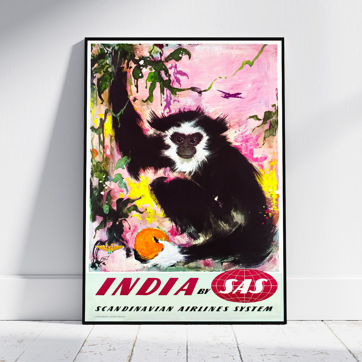 A framed vintage-style travel poster leaning against a white wall on a light floor. The artwork shows a black-and-white primate in a pink, yellow, and green illustrated scene, with the text 'INDIA by SAS' and 'SCANDINAVIAN AIRLINES SYSTEM' at the bottom.
