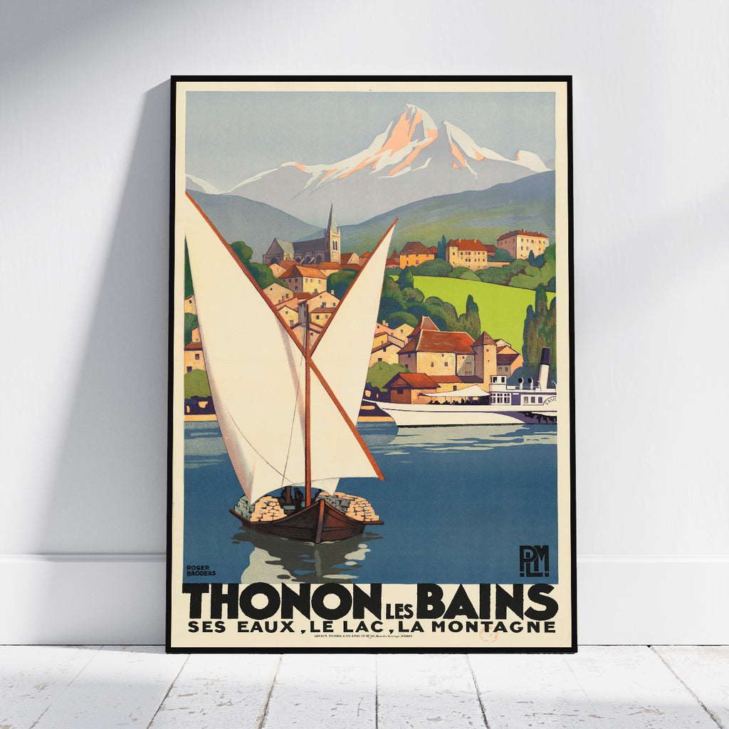 A framed vintage travel poster leaning against a white wall on a light floor. The poster shows a sailboat on blue water with a coastal town, green hills, and a snow-capped mountain in the background.