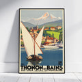 A framed vintage travel poster leaning against a white wall on a light floor. The poster shows a sailboat on blue water with a coastal town, green hills, and a snow-capped mountain in the background.
