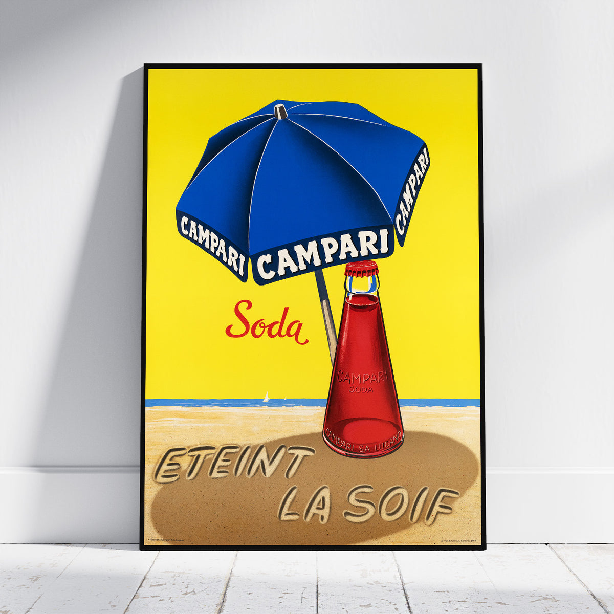 A framed vintage-style poster leaning against a white wall on a light floor. The poster shows a blue beach umbrella with CAMPARI branding, a red soda bottle, and the text 'ETEINT LA SOIF' on a yellow background.