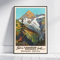 A framed vintage-style travel poster leaning against a white wall on a light floor. The artwork shows a mountain landscape with snow, green and brown slopes, trees, and a small rider in the lower left.