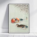 A framed Japanese-style art print depicting two ducks on a pale green water surface with reeds at the top, leaning against a white wall on a light floor.