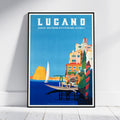 A framed vintage-style travel poster for Lugano, Switzerland, leaning against a white wall on a light wood floor. The poster features a bright blue sky and sea, colorful coastal buildings, a sailboat, and bold white text reading 'LUGANO' with smaller text 'SWISS SOUTHERN SWITZERLAND - SCHWEIZ'.