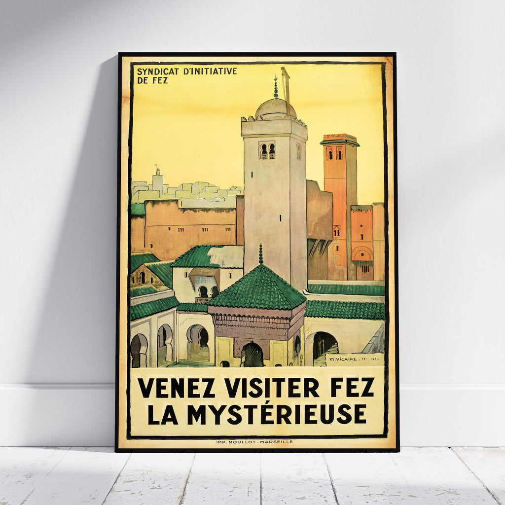 A framed vintage travel poster of Fez, Morocco, leaning against a white wall on a light wooden floor. The poster features a yellow sky and architectural details of the city with the French text 'VENEZ VISITER FEZ LA MYSTÉRIEUSE' at the bottom.